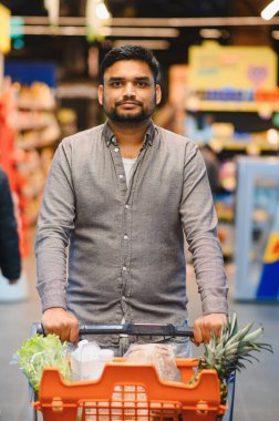 Genç adam alışveriş yapıyor, taze ürünlerle dolu bir arabayı parlayan bir markette ananas ve marul gibi itiyor.