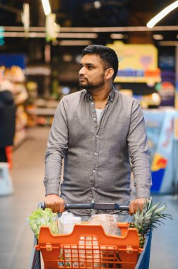 Modern bir markette içi taze sebze, ananas ve diğer yiyecek malzemeleriyle dolu bir alışveriş arabasını iten adam.