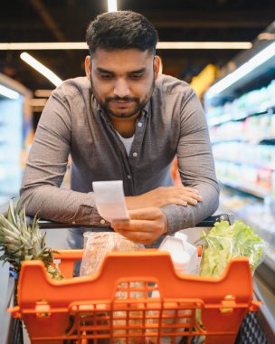 Elinde alışveriş listesi olan bir adam, parlak bir süpermarket koridorunda içi taze yiyecek dolu portakal tezgahına yaslanmış.