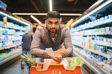 Süpermarket koridorunda alışveriş arabasıyla dolu uzun bir fişe bakan bir adam. Artan gıda maliyetlerine odaklan
