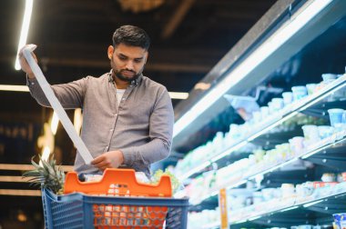 Süpermarkette bir adam, soğutulmuş rafların yanında yiyecek dolu bir alışveriş arabasını iterken uzun bir market listesini inceliyor.