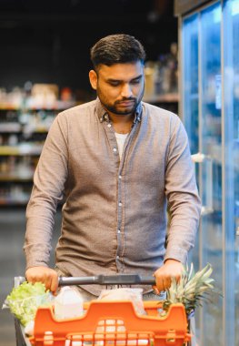 Taze ürünler ve gerekli malzemelerle dolu bir alışveriş arabasını iten adam, bir manavın koridorunda geziniyor.