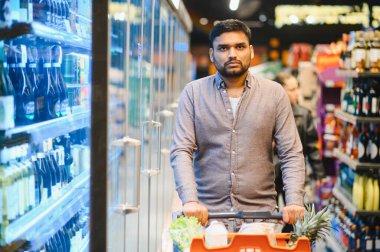 Market alışverişi yapan, dolu bir arabayı soğutulmuş içecek vitrinlerinin ve ürün raflarının önünden iten bir adam.