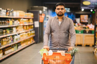 Modern bir süpermarket koridorunda alışveriş arabası süren bir adam, günlük ihtiyaçlar ve yemekler için taze ürünler ve bakkaliye seçiyor.