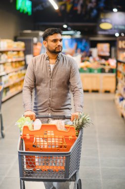 Taze ürün ve paketlerle dolu bir alışveriş arabasını iten bir adam süpermarket koridorunda yürüyor.