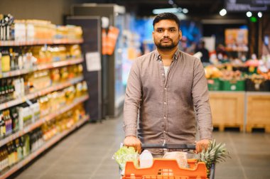 Genç bir adam markette yiyecek dolu bir alışveriş arabasını itiyor, günlük ihtiyaçlara ve tüketime odaklanıyor.