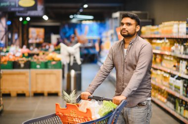 Genç adam, içi taze yiyecek dolu bir alışveriş arabasını itiyor. Parlak bir markette koridorlarda geziniyor.