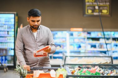 Market koridorunda içi taze ürünlerle dolu bir arabayı iterken elinde ambalaj malzemesi tutan genç bir adam.