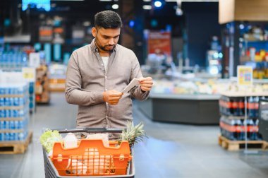 Süpermarket koridorunda alışveriş arabasını iten adam alışveriş listesini dikkatlice kontrol ediyor.