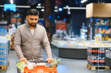 Modern bir market koridorunda içi taze meyve, ananas ve diğer yiyecek malzemeleriyle dolu alışveriş arabası satan bir adam.