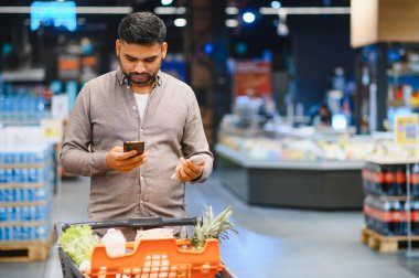 Süpermarkette alışveriş yapan bir adam, akıllı telefondan dijital alışveriş listesini kontrol ederken aynı zamanda içi ürünlerle dolu bir arabayı itiyor.