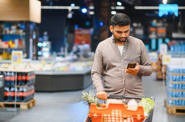 Genç bir adam market arabasıyla alışveriş yapıyor, cep telefonundaki eşyaları ve fiyatları modern bir süpermarket koridorunda karşılaştırıyor.