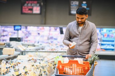 Genç adam bir parça peynir tutuyor, modern bir markette soğutulmuş bir vitrinin yanında duruyor.