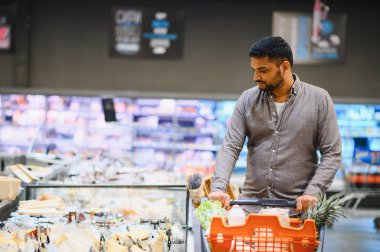 Genç adam marketin koridorunda içi taze ürün, mandıra ve diğer eşyalarla dolu bir alışveriş arabasını itiyor.