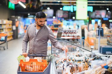 Market alışverişi yapan genç bir adam, modern bir süpermarket koridorundaki soğutuculu vitrinlerden taze ürünler seçiyor.