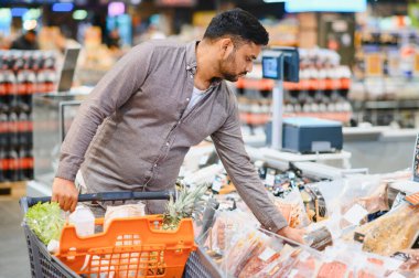 Süpermarketten alışveriş yapan genç adam, market koridorunda alışveriş yapıyor, el arabasını itiyor ve modern marketten taze meyve ve sebze seçiyor.