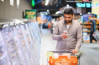 Adam yiyecek alıyor, ürün etiketini dikkatlice okuyor ve modern bir markette alışveriş arabasını itiyor.