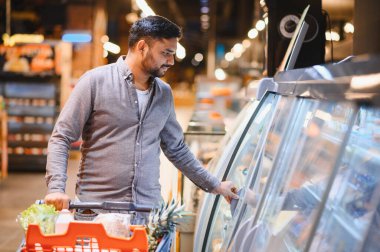 Market alışverişi yapan genç bir adam, canlı bir süpermarket koridorunda alışveriş arabasıyla soğutulmuş bir kutu açıyor.