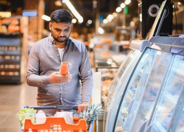 Bir süpermarket koridorunda alışveriş arabasını iterken paketlenmiş bir ürünü inceleyen bir adam, satın almadan önce ürünleri karşılaştırıyor.