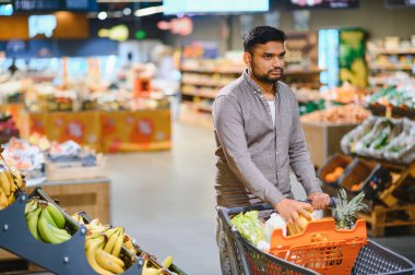 Taze meyve ve sebzelerle dolu bir alışveriş arabasını iten adam, modern bir marketten eşyalar seçiyor.