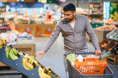Adam marketten taze muz seçiyor, içi sağlıklı yiyeceklerle dolu bir arabayı itiyor. Meyve alan tüketici