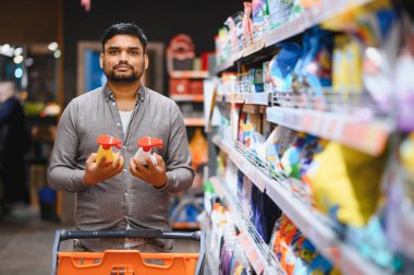 Markette alışveriş yapan genç bir adam, iki sprey temizlik malzemesini ev içi kullanım için karşılaştırıyor.