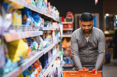 Market alışverişi yapan, perakende mağazasında çeşitli ürünlerle dolu rafları karıştıran genç bir adam. Günlük tüketim