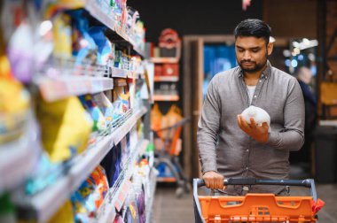 Elinde ürün şişesi olan bir adam, alışveriş arabasını kalabalık bir süpermarket koridorunda iterken etiketi okuyor.