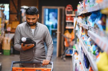 Bir şişe sıvı deterjan tutan bir adam, yoğun bir mağaza koridorunda alışveriş arabasını iterken ürünleri dikkatle karşılaştırıyor.