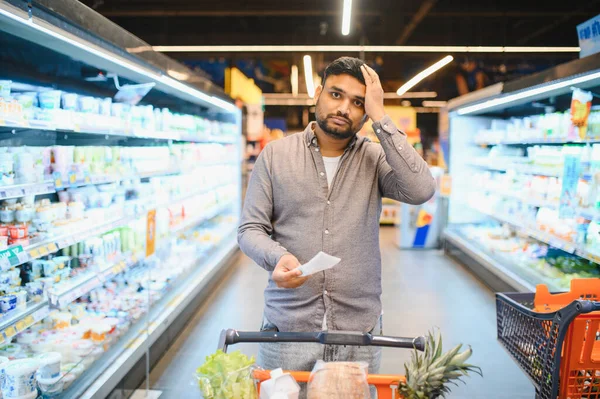 Adam elinde uzun bir makbuz tutarken stresli ve şaşkın hissediyor, süpermarkette yiyecek alışverişi yapıyor, enflasyon yaşıyor.