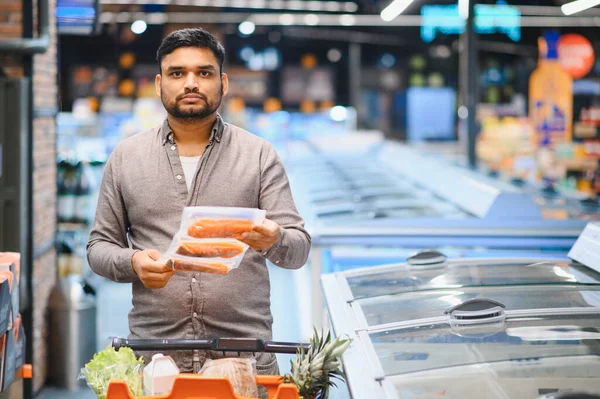 Modern bir süpermarketin dondurucu koridorunda alışveriş yaparken paketlenmiş somon filetosu tutan adam, tüketicilik...