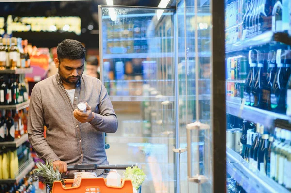 Market koridorunda soğutmalı içeceklerle alışveriş arabası tutan bir adam, ürün etiketini dikkatle kontrol ediyor.