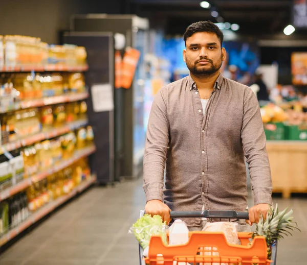 Market alışverişi yapan, alışveriş arabasını koridorda iten, kameraya bakan genç bir adam. Tüketim ve perakende kavramı