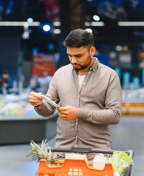 Alışveriş listesini kontrol eden bir adam süpermarkette alışveriş arabasıyla dikkatli alışverişe odaklanıyor.