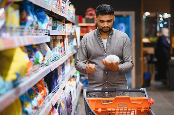 Asyalı bir adam deterjan alışverişi yapıyor, markette ürün seçeneklerini karşılaştırıyor, satın alma kararı veriyor.