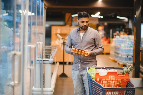 Müşteri bir karton yumurtayı elinde tutarken market koridorunda içi yiyecek dolu bir alışveriş arabasını itiyor.