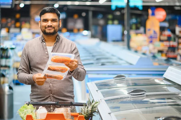 Bir markette paketlenmiş somon tutan genç adam sağlıklı yiyecekler için taze yiyecek ve malzeme alıyor.