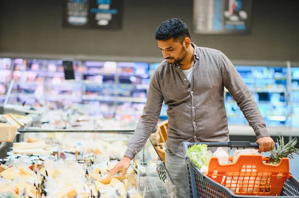 Dondurulmuş bir vitrinden taze ürünler seçen bir adam süpermarket koridorunda içi yiyecek dolu bir alışveriş arabasını itiyor.
