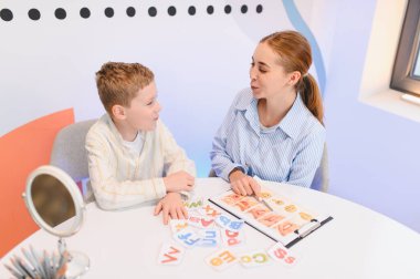 Genç bir çocuk konuşma terapistiyle bire bir ders alıyor, alfabe kartları ve ayna kullanarak dil telaffuzuna odaklanıyor.