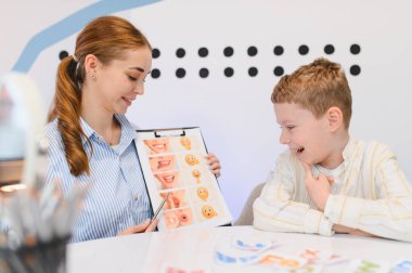 Konuşma terapisti genç bir çocuğa dil gelişiminde yardım ediyor, bir seans sırasında panoda ifade egzersizleri gösteriyor.