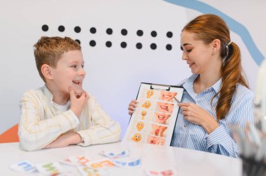 Kadın konuşma terapisti, iletişim becerilerini geliştirmek için yüz ifadesi grafiklerini kullanarak çocuğa konuşma dersi vererek yardımcı oluyor.