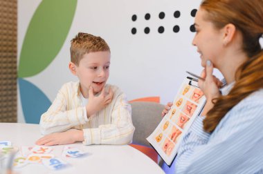 Genç bir çocuk, seans sırasında görsel çizelgeler ve öğretim materyalleri kullanarak kadın bir terapistten konuşma dili öğreniyor.