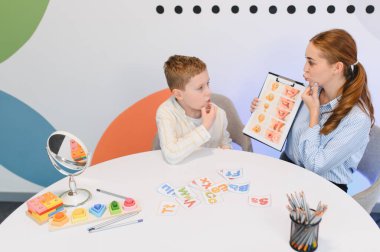 Konuşma terapisti ve genç bir çocuk, renkli eğitim materyalleri ve ayna kullanarak konuşma eklemleri üzerinde çalışıyor.