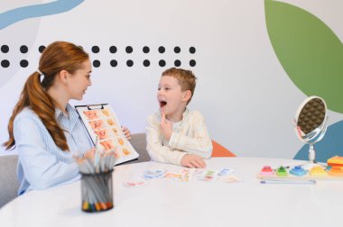 Konuşma terapisti genç bir çocukla çalışıyor, dil geliştirme oturumunda ona konuşma konusunda yardım ediyor. Oral motor alıştırması için görsel araçlar kullanılıyor