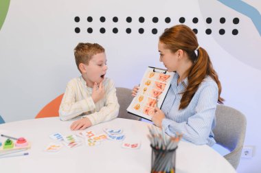 Konuşma terapisti bir ders sırasında genç bir çocukla çalışıyor, görsel bir çizelge kullanarak konuşma ve fonetik eğitimi almasına yardım ediyor.