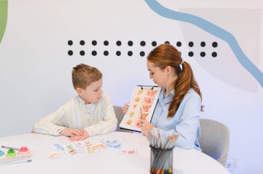 Konuşma terapisti genç bir çocukla çalışıyor, farklı ağız şekilleri ve duyguları olan kartları göstererek konuşma ve konuşma yetisini geliştirmesine yardım ediyor.