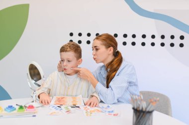 Konuşma terapisti genç bir oğlanla çalışıyor, eklem egzersizleri ve ayna kullanarak oral motor beceri eğitiminde ona yol gösteriyor.