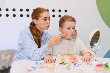 Yetişkin konuşma terapisti, konuşma ve telaffuz egzersizlerinde küçük bir çocuğa yardımcı oluyor, seans sırasında aynaya bakıyor.
