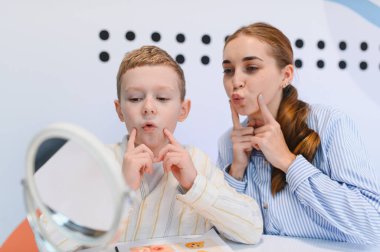 Çocuk, konuşma terapistiyle aynanın karşısında telaffuz ve iletişim becerilerine odaklanmış bir şekilde konuşma egzersizleri yapıyor.