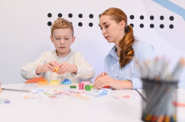 Genç bir çocuk renkli ahşap blokları istiflemeye konsantre olurken, bir öğretmen onun gelişimini izliyor, erken eğitim ve gelişimi destekliyor.